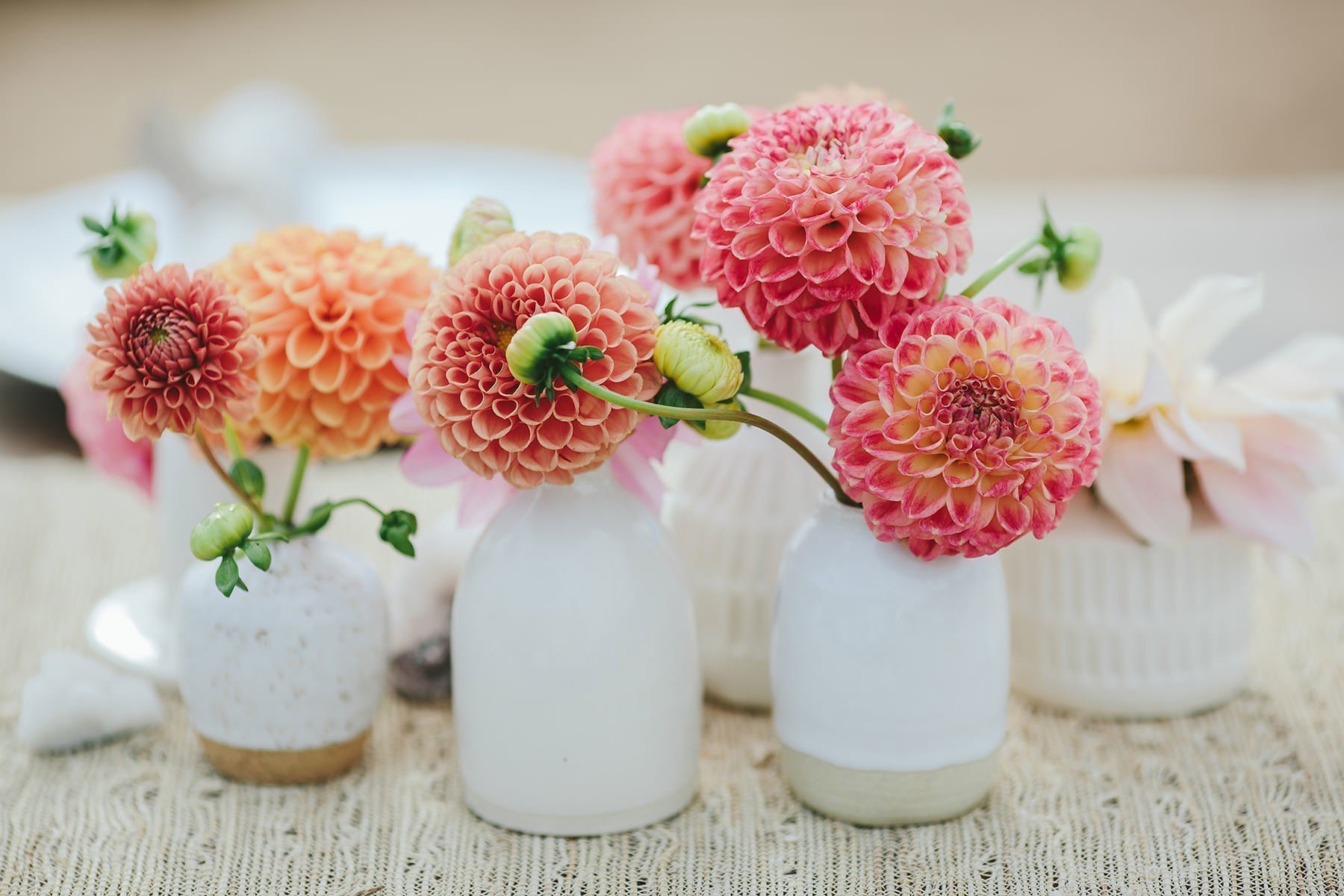 Small handmade white ceramic bud vases holding pink and coral dahlias on a woven table runner